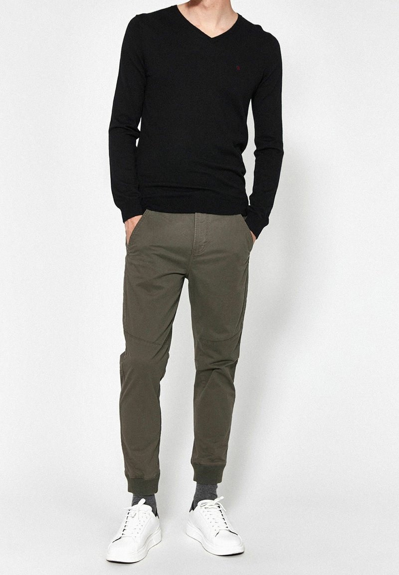 Černý svetr s výstřihem do V, olivově zelené zúžené kalhoty, bílé tenisky. Kalhoty mají boční kapsy, žebrované manžety. Texturovaná látka po celém povrchu.