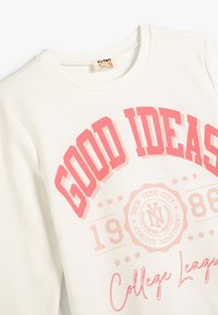 Sudadera de color crema claro con un atrevido texto gráfico rojo "BUENAS IDEAS" y "Liga Universitaria", con detalles de logotipo, textura suave y cuello acanalado.