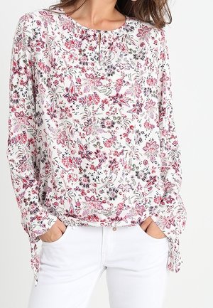 Mujer con una blusa blanca de manga larga con patrones florales en rosa y rojo, combinada con pantalones blancos, con las manos en los bolsillos.