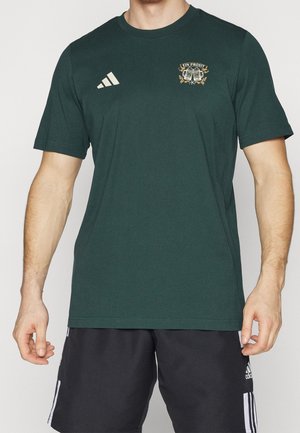 Mężczyzna w ciemnozielonej koszulce Adidas z logo „Ein Prosit” oraz czarnych spodenkach Adidas z białymi paskami i logo po bokach.