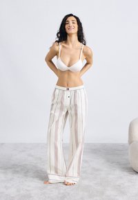 Λευκό σουτιέν συνδυασμένο με φαρδιά, βαμβακερή πιτζάμα με ρίγες σε κόκκινες και μπλε γραμμές και ελαστική μέση. Γυμνά πόδια πάνω σε ανοιχτό γκρι χαλί.