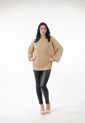 Donna in piedi con le mani sui fianchi, che indossa un maglione beige a maglia grossa, pantaloni in pelle nera e tacchi bianchi aperti sulle dita dei piedi, su sfondo bianco.