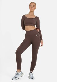 Smeđi kratki crop top s dugim rukavima i tajice visokog struka od teksturirane tkanine. Sadrži bijeli logo na lijevoj strani prsnog koša i sa strane hlača.