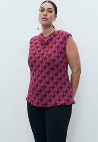 Red and purple patterned sleeveless blouse with a draped neckline and a relaxed fit, paired with black pants.