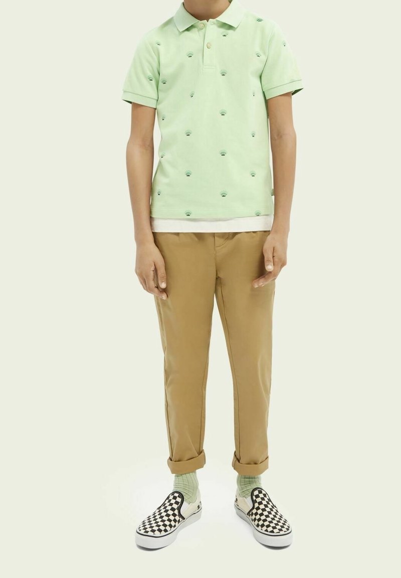 Personne portant un polo vert clair avec de petits motifs verts, un pantalon beige retroussé, des chaussettes vertes et des chaussures slip-on à carreaux noir et blanc.