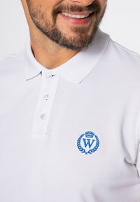 Biała koszulka polo o fakturowanej tkaninie, z niebieskim haftowanym logo z koroną i laurowym wzorem oraz zapięciem na trzy guziki.