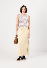 Débardeur rayé blanc et marron, associé à une longue jupe jaune avec une fente sur le côté, et des baskets noires avec des rayures blanches. Sac à main marron.
