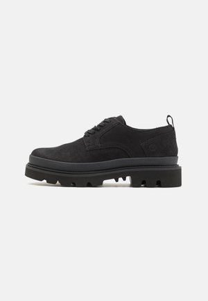 Schwarze Wildlederschuhe mit einer dicken schwarzen Gummisohle, ausgestattet mit Schnürverschluss und einem Zugschlaufe an der Ferse. Strukturierte Oberfläche und robustes Design.