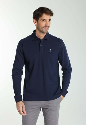 Homme portant un polo bleu marine à manches longues et un pantalon gris clair, debout avec une main dans la poche, regardant sur le côté, fond neutre.