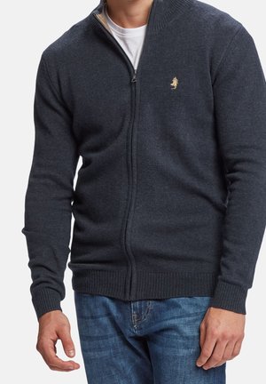 Man draagt een donkergrijs gebreid vest met rits en een klein logo op de borst, gecombineerd met een blauwe spijkerbroek, staand tegen een effen achtergrond.