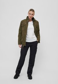 Chaqueta militar en verde oliva con parches y bolsillos con cremallera; combinada con una camiseta blanca y pantalones cargo negros. Zapatos negros. Fondo minimalista.