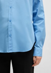 Camicia azzurro chiaro con tessuto liscio, colletto a punta e maniche lunghe. Orlo curvo e dettaglio del polsino con un bottone.
