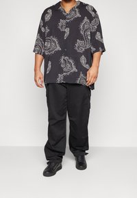 Mies, joka on pukeutunut mustaan paitaan, jossa on valkoisia paisley-kuvioita, mustiin cargo-housuihin ja mustiin lenkkareihin, seisoo yksinkertaista taustaa vasten.