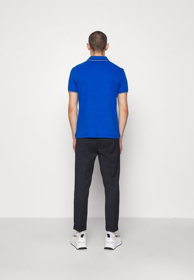 Μπλε πόλο μπλούζα με αντίθεση στο γιακά και το τελείωμα των μανικιών, σε συνδυασμό με σκούρες παντελόνες. Το μοντέλο φοράει λευκά sneakers. Απλό φόντο.