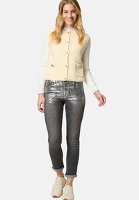 Cremfarbener gerippter Pullover mit Knöpfen und Taschen, kombiniert mit glänzenden silbernen Skinny Jeans und cremefarbenen Sneakers, vor einem neutralen Hintergrund.