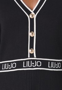 Pull structuré noir avec un col en V, des boutons dorés, des bordures blanches et une ceinture avec logo en texture côtelée.