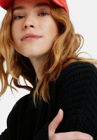 Black knit sweater with a textured pattern, featuring long sleeves. A red baseball cap is worn. The model has wavy, shoulder-length hair.