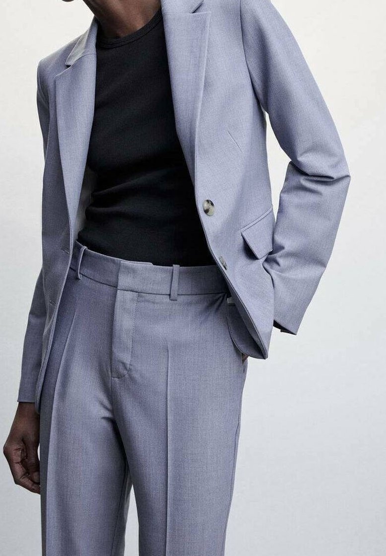Personne portant un costume gris clair avec un pantalon sur-mesure et une veste par-dessus une chemise noire ajustée, avec une main dans la poche, sur un fond uni.