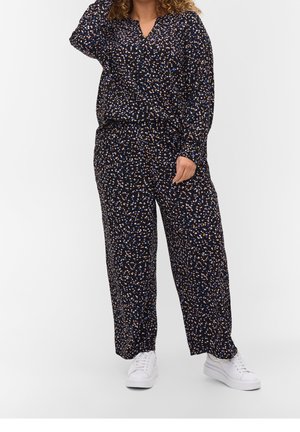 Person iført sort langærmet top og bukser med flerfarvet lille prikmønster, kombineret med hvide sneakers, stående på hvid baggrund.