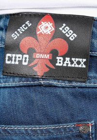 Étiquette en cuir noir avec un design de fleur-de-lis rouge, texte blanc. Tissu en jean bleu, avec quincaillerie en métal et détails de couture visibles.