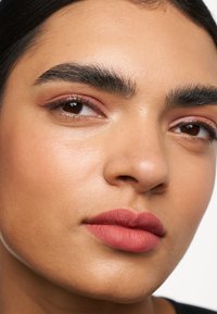 Primer plano de un rostro con cabello oscuro, cejas definidas y lápiz labial rosa mate. Piel suave con un ligero tono cálido y acentos de sombra de ojos sutiles.