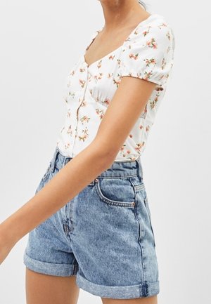 Kobieta ubrana w białą, kwiecistą bluzkę z krótkim rękawem na guziki oraz wysokiej talii, podwinięte szorty jeansowe na jednolitym tle.