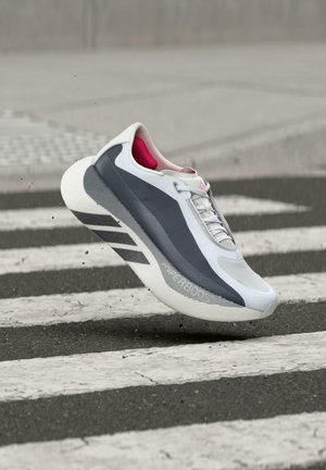 White and gray athletic shoe with pink interior mid-air above a black and white striped crosswalk on asphalt street.