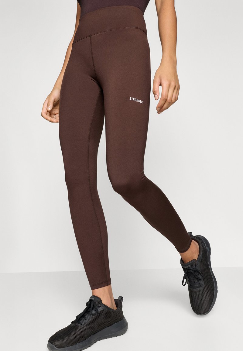 Bruine legging van rekbaar materiaal met een hoge taille. Bevat een wit "STRONGER" logo en heeft een gladde, aansluitende pasvorm. Zwarte sportschoenen zichtbaar.