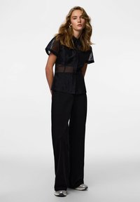 Črna majica s kratkimi rokavi z prosojnim prekrivnim slojem in zapenjanjem na gumbe, v kombinaciji z ohlapnimi črnimi hlačami in srebrnimi supergami.