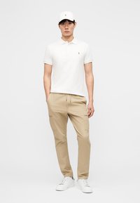 Fehér pólóingben, fehér sapkában, bézs cargo nadrágban és fehér tornacipőben álló férfi, kezeit zsebében tartva, fehér háttér előtt.