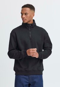 Sweat à zip noir avec col montant, poignets côtelés et texture douce, associé à un jean en denim bleu.