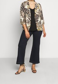 Jakke med blomstermønster i creme, kombineret med en sort v-hals top, vide mørke denimbukser og brune sandaler med remme.