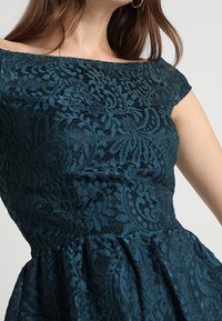 Robe en dentelle bleu sarcelle avec encolure bardot, motifs floraux complexes, corsage structuré et jupe évasée.