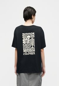 Άτομο με κοντά σκούρα μαλλιά που φοράει μαύρο μπλουζάκι με στυλιζαρισμένα γραφικά ήλιου, λουλουδιού και δέντρου και το κείμενο "COLUMBIA" στην πλάτη.