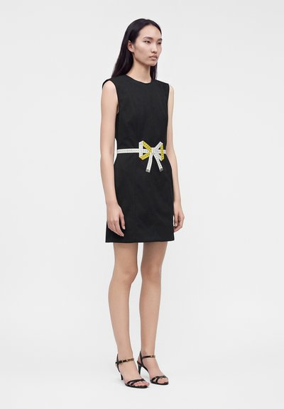 Femme portant une robe noire sans manches avec un ruban à mesurer blanc et jaune noué en nœud autour de la taille, associée à des sandales noires à talons hauts.