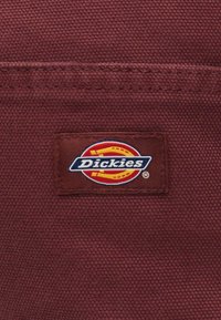 Tecido texturizado vermelho escuro com um rótulo retangular cosido apresentando o logo da Dickies em vermelho, amarelo, branco e azul-marinho.