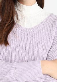 Pull en maille violet clair avec un col en V et des poignets côtelés, superposé sur un col roulé blanc côtelé, doté d'un tissu lâche et texturé.