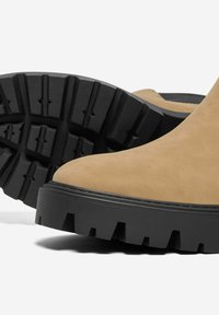Botas de tobillo de ante en color marrón claro con suela de goma negra gruesa que presenta patrones de relieve profundos y un diseño de puntera redonda.