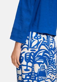 Royal blue linen blouse with long sleeves, paired with beige pants featuring a bold blue abstract print of palm trees and wine glasses.