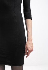Gros plan sur le bras droit et la main d'une personne portant une robe noire ajustée à manches longues jusqu'au coude, sur fond blanc uni.