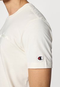 Κρεμ κοντομάνικη μπλούζα με στρογγυλή λαιμόκοψη, απαλό ύφασμα και κεντημένο λογότυπο στο αριστερό μανίκι.