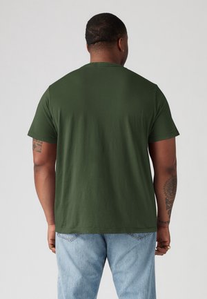 Mand med tatoveringer iført en ensfarvet mørkegrøn kortærmet T-shirt og lyseblå jeans, vist bagfra mod en ensfarvet baggrund.