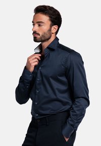 Marineblauwe blouse met lange mouwen van een gladde stof, voorzien van knopen en een patroon op de kraag. Getailleerde pasvorm met schouderaccenten.