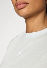 T-shirt de sport gris clair avec une texture respirante et un col côtelé blanc. Présente un logo Jordan blanc sur la zone gauche de la poitrine.