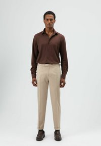 Bruine blouse met lange mouwen en knopen, gecombineerd met lichtbeige broek. De broek heeft een nauwsluitende pasvorm en zijzakken. Bruine loafers maken de look compleet.