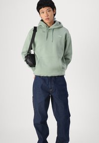 Sudadera verde con cordón, bolsillo canguro y logo blanco, combinada con pantalones de mezclilla azul que tienen costuras visibles y un bolsillo lateral.