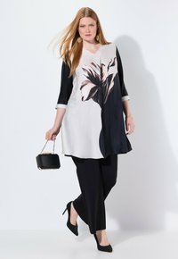 Zwart-witte tuniek met een bloemenpatroon, gecombineerd met zwarte broeken. De outfit heeft lange mouwen en een v-hals, aangevuld met zwarte hakken.