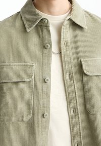 Chaqueta de camisa de pana en verde claro, con cuello, cierre de botones en la parte delantera y dos bolsillos en el pecho. Tejido texturizado con estrías verticales.