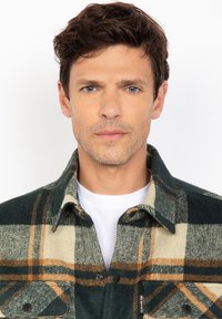 Primo piano di un uomo con capelli corti castani e occhi blu che indossa una giacca a quadri verde, beige e arancione sopra una camicia bianca su sfondo bianco.