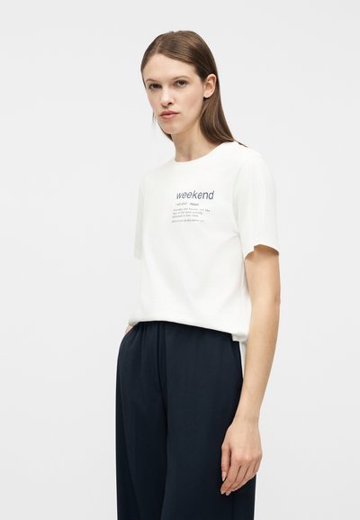 Νεαρή γυναίκα με ίσια καστανά μαλλιά, φορούσα λευκό t-shirt με το γράφημα "weekend" και άνετο μαύρο παντελόνι, στέκεται μπροστά σε ένα απλό φωτεινό φόντο.